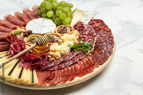 Antipasto platter cold meat plate with grissini bread sticks, prosciutto, slices ham, grapes, jamon ham, beef jerky, chorizo salami, fuet, pate and honey. Camembert goat cheese on wooden cutting board
