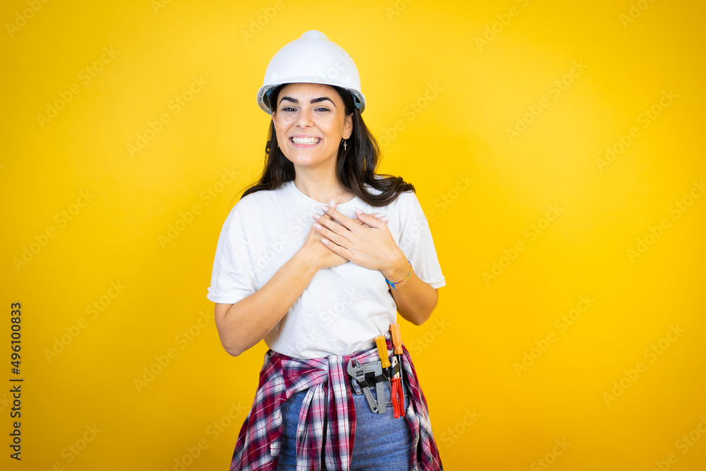 Young caucasian woman wearing hardhat and builder clothes over isolated yellow background smiling with her hands on her chest and grateful gesture on her face. Young caucasian woman wearing hardhat and builder clothes over isolated yellow background smiling with her hands on her chest and grateful gesture on her face.