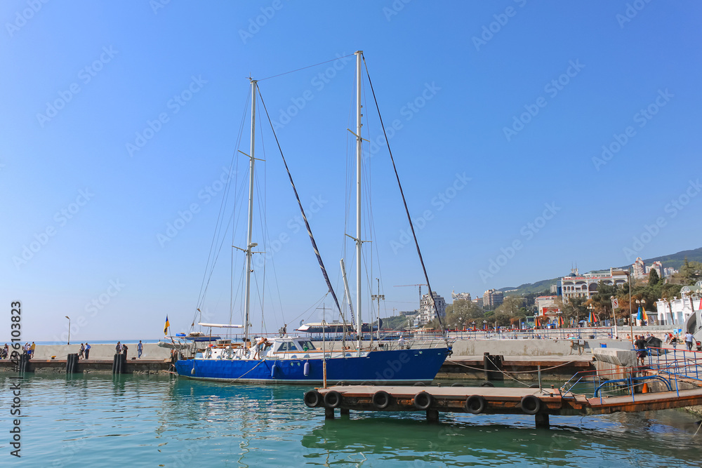 A blue yacht with high masts stands in the port. In the background is a blue sky, city and mountains. Yalta, Crimea.