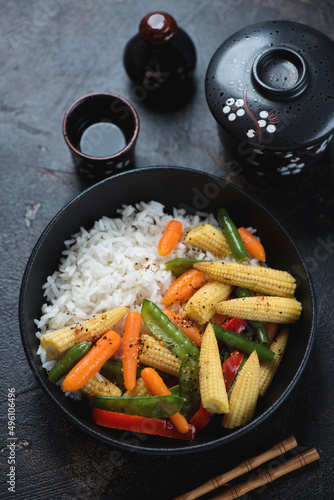 Black bowl with vegetable stir-fry and rice, vertical shot on a dark-brown stone background, elevated view