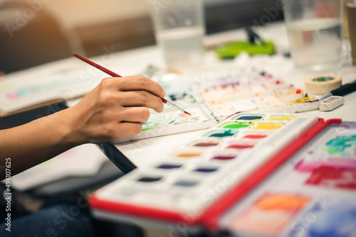 A woman's hand holding a brush to paint watercolors placed on the table.