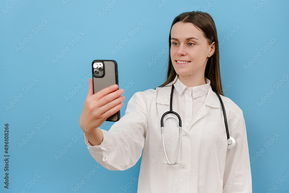 Smiling young female doctor holding phone talk to patient make telemedicine online conference video call, isolated on blue wall. Gp remote consultation telemedicine virtual mobile health care chat app Smiling young female doctor holding phone talk to patient make telemedicine online conference video call, isolated on blue wall. Gp remote consultation telemedicine virtual mobile health care chat app