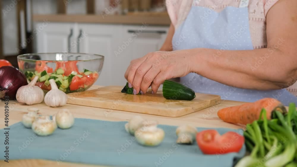 Closeup of Senior Woman in Apron Chopping Cucumbers on the Wooden Board in the Kitchen. Slow Motion. Cooking and Healthy Vegan Eating Concept Closeup of Senior Woman in Apron Chopping Cucumbers on the Wooden Board in the Kitchen. Slow Motion. Cooking and Healthy Vegan Eating Concept