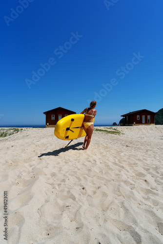 Back view relaxed female surfer with tanned skin in yellow swimsuit standing on beach with surfboard photo