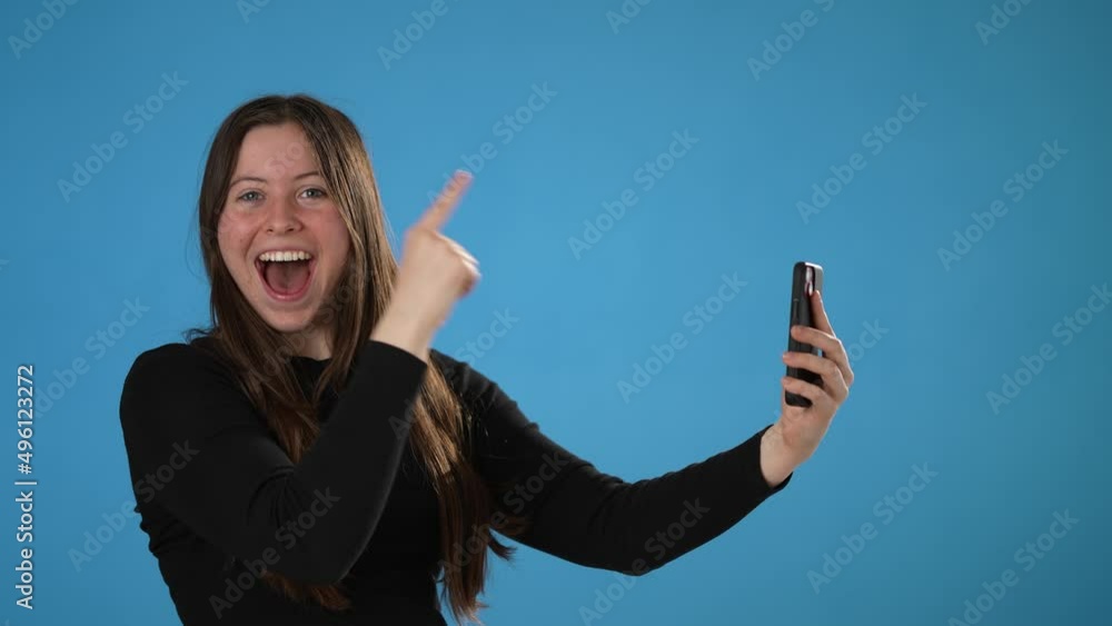 Surprised young woman 20s posing isolated on blue background studio. Using mobile cell phone typing sms message pointing index finger to copy space say wow Surprised young woman 20s posing isolated on blue background studio. Using mobile cell phone typing sms message pointing index finger to copy space say wow