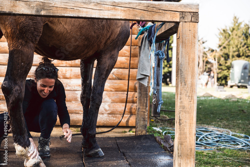 Female owner with a big smile taking care of brown horse hoof, washing legs with water and shampoo in outdoors stable on animal farm