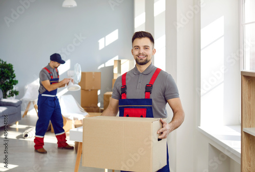 Smiling young male deliveryman or mover hold cardboard box pack belongings in client office or home. Happy man worker from removal or deliver company unload packages. Moving and relocation. photo