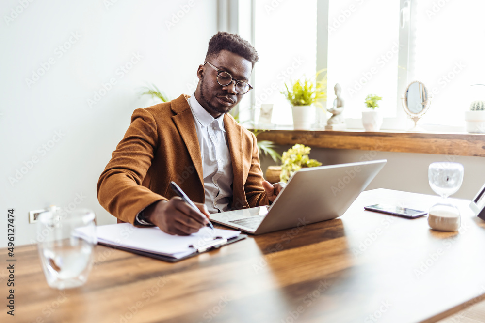 custom made wallpaper toronto digitalConcentrated African man in glasses sit at desk work on computer make notes. Focused serious male worker or freelancer busy at home office watch webinar on laptop, handwrite on paper.