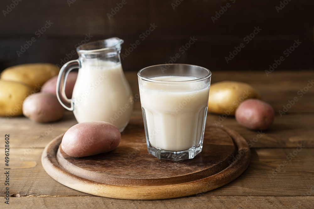 Vegan potato milk in glass and milk jar on cutting board with raw potatoes aside. Wooden background. Rustic style Vegan potato milk in glass and milk jar on cutting board with raw potatoes aside. Wooden background. Rustic style