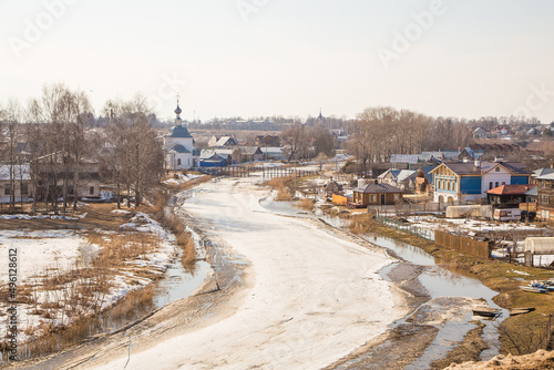 A crooked, frozen river stretching into the distance, wooden houses on the shore. Spring, snow melts, puddles and dry grass all around. Day, cloudy weather, soft warm light.