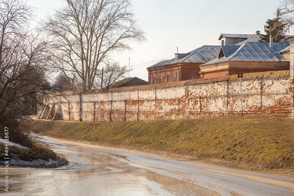 A wet road going down, an old brick wall and roofs of houses on the hill behind. Spring, snow melts, dry grass all around, small snowdrifts and puddles. Day, cloudy weather, soft warm light.