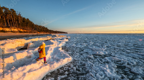 Little girl in yellow jacket walks on snow on sea shore.