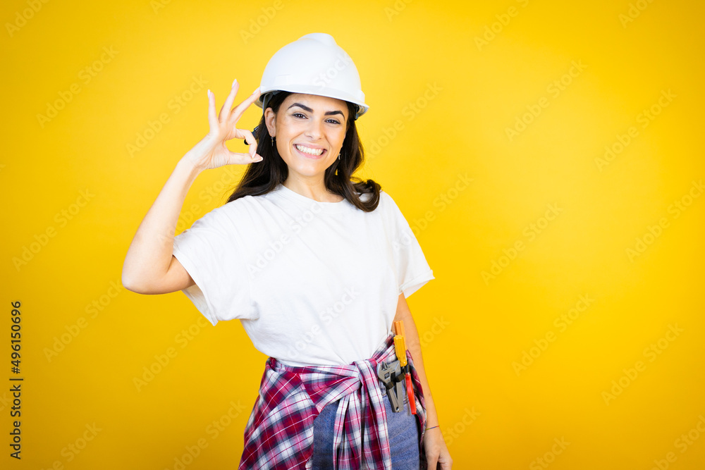 Young caucasian woman wearing hardhat and builder clothes over isolated yellow background doing ok sign with fingers and smiling, excellent symbol