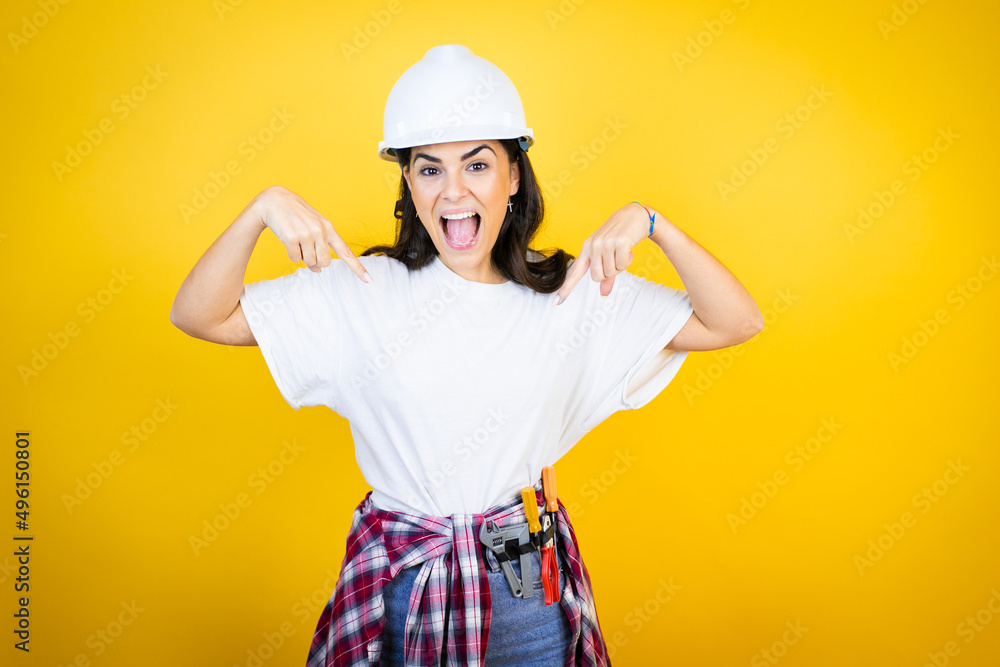 Young caucasian woman wearing hardhat and builder clothes over isolated yellow background looking confident with smile on face, pointing oneself with fingers proud and happy.