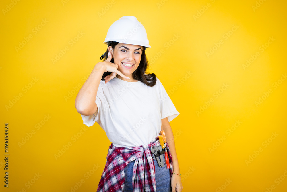 Young caucasian woman wearing hardhat and builder clothes over isolated yellow background smiling doing phone gesture with hand and fingers like talking on the telephone Young caucasian woman wearing hardhat and builder clothes over isolated yellow background smiling doing phone gesture with hand and fingers like talking on the telephone