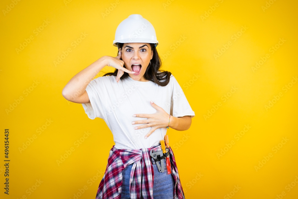 Young caucasian woman wearing hardhat and builder clothes over isolated yellow background confused doing phone gesture with hand and fingers like talking on the telephone Young caucasian woman wearing hardhat and builder clothes over isolated yellow background confused doing phone gesture with hand and fingers like talking on the telephone