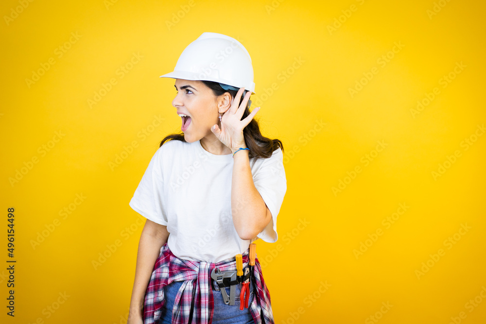 Young caucasian woman wearing hardhat and builder clothes over isolated yellow background surprised with hand over ear listening an hearing to rumor or gossip