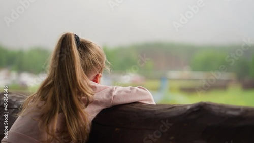 Adorable little girl leans on log handrail and watches helicopter starting engine from terrace of resort cafe on rainy day slow motion photo