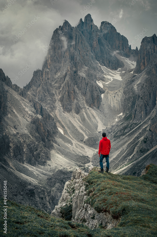 Man with red jacket in front of mountain peaks in the Dolomite Alps in South Tyrol with dramatic cloudy sky, Three Peaks Nature Reserve, Italy.