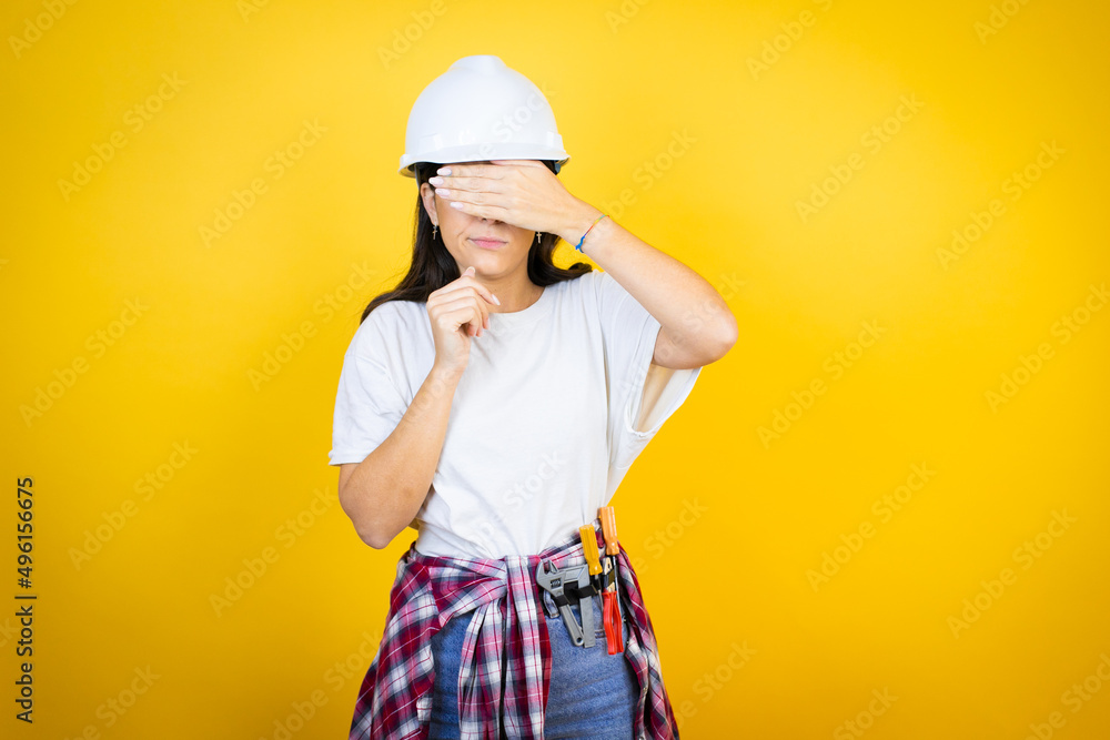 Young caucasian woman wearing hardhat and builder clothes over isolated yellow background serious and covering her eyes with her hand