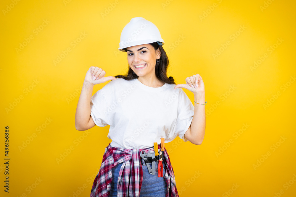 Young caucasian woman wearing hardhat and builder clothes over isolated yellow background looking confident with smile on face, pointing oneself with fingers proud and happy. Young caucasian woman wearing hardhat and builder clothes over isolated yellow background looking confident with smile on face, pointing oneself with fingers proud and happy.