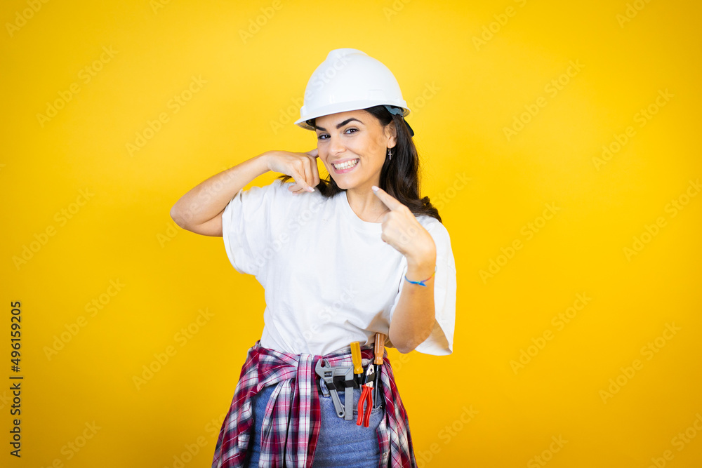 Young caucasian woman wearing hardhat and builder clothes over isolated yellow background doing the “call me” gesture with her hands. Young caucasian woman wearing hardhat and builder clothes over isolated yellow background doing the “call me” gesture with her hands.