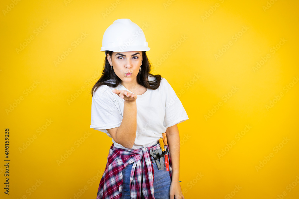 Young caucasian woman wearing hardhat and builder clothes over isolated yellow background looking at the camera blowing a kiss with hand on air being lovely and sexy. Love expression.