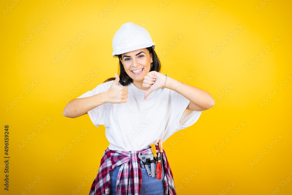 Young caucasian woman wearing hardhat and builder clothes over isolated yellow background Doing thumbs up and down, disagreement and agreement expression. Crazy conflict