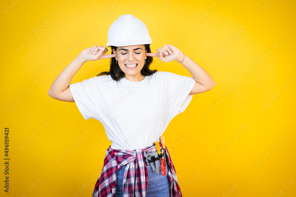 Young caucasian woman wearing hardhat and builder clothes over isolated yellow background covering ears with fingers with annoyed expression for the noise of loud music. Deaf concept.