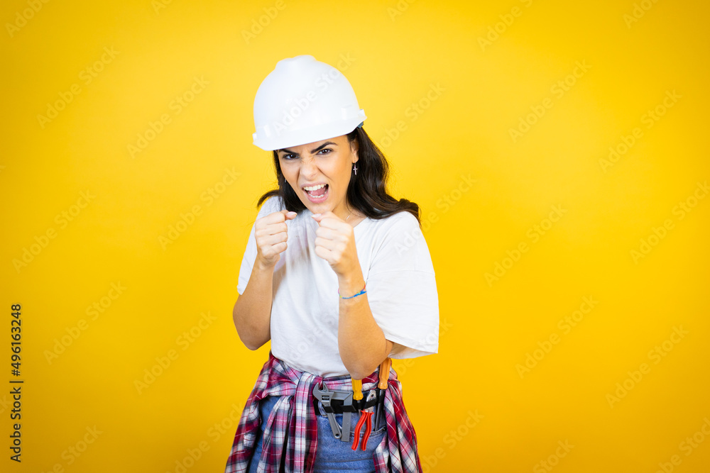 Young caucasian woman wearing hardhat and builder clothes over isolated yellow background Punching fist to fight, aggressive and angry attack, threat and violence Young caucasian woman wearing hardhat and builder clothes over isolated yellow background Punching fist to fight, aggressive and angry attack, threat and violence