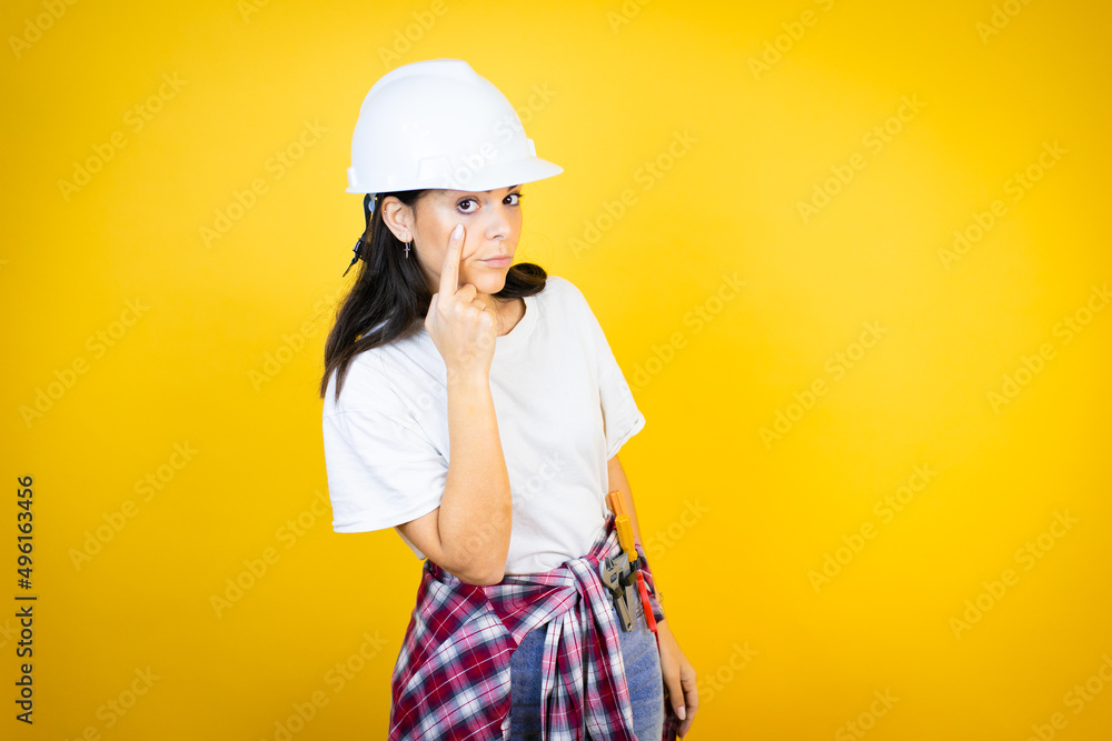 Young caucasian woman wearing hardhat and builder clothes over isolated yellow background Pointing to the eye watching you gesture, suspicious expression Young caucasian woman wearing hardhat and builder clothes over isolated yellow background Pointing to the eye watching you gesture, suspicious expression