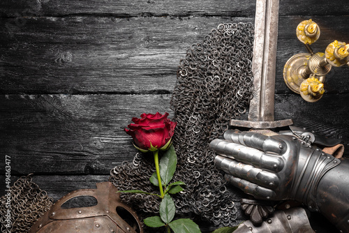 Φωτογραφία Knight sword, red rose flower and iron helm on the black woorden flat lay table background