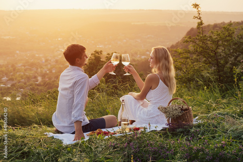 Summer picnic outdoors. Romantic happy couple is toasting glasses with wine and smiling. Romantic picnic with mountain view at sunset.