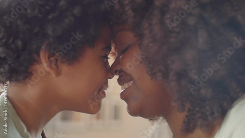 Close up shot of affectionate African American mother and happy little son smiling and rubbing their noses in room filled with sunlight photo