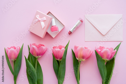 top view of row with blooming tulips near gift box with nail polish near lipstick and envelope on pink.