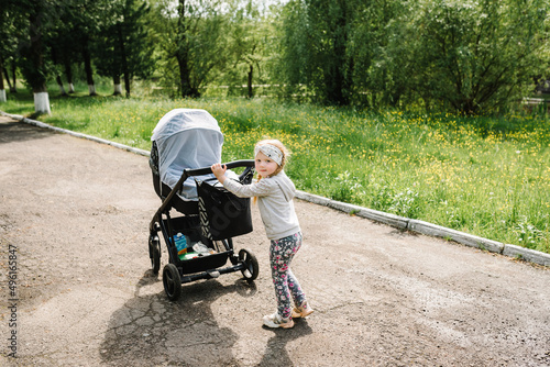 A little girl goes and carries a stroller with her little brother or sister in the park. Walking in the summer. © Serhii