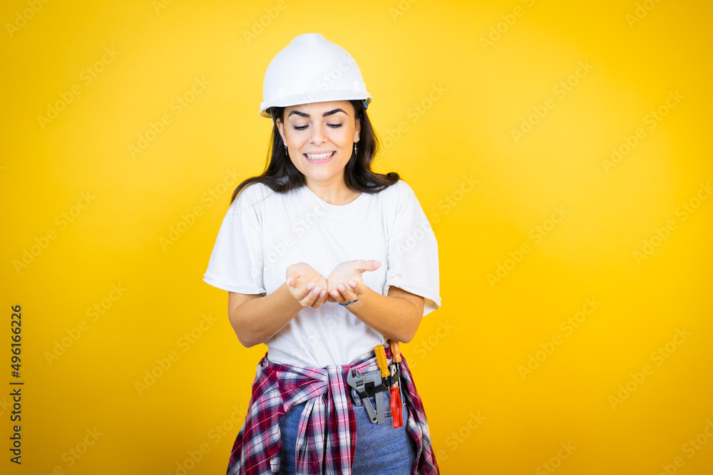 Young caucasian woman wearing hardhat and builder clothes over isolated yellow background Smiling with hands palms together receiving or giving gesture. Hold and protection Young caucasian woman wearing hardhat and builder clothes over isolated yellow background Smiling with hands palms together receiving or giving gesture. Hold and protection