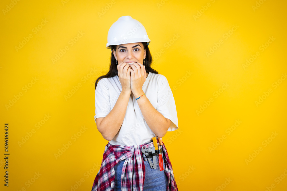 Young caucasian woman wearing hardhat and builder clothes over isolated yellow background looking stressed and nervous with hands on mouth biting nails. anxiety problem. Young caucasian woman wearing hardhat and builder clothes over isolated yellow background looking stressed and nervous with hands on mouth biting nails. anxiety problem.