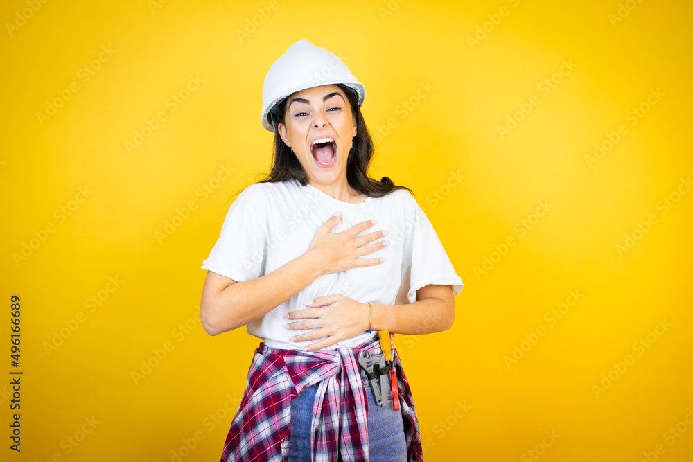 Young caucasian woman wearing hardhat and builder clothes over isolated yellow background laughs happily and has fun keeping hands on stomach.
