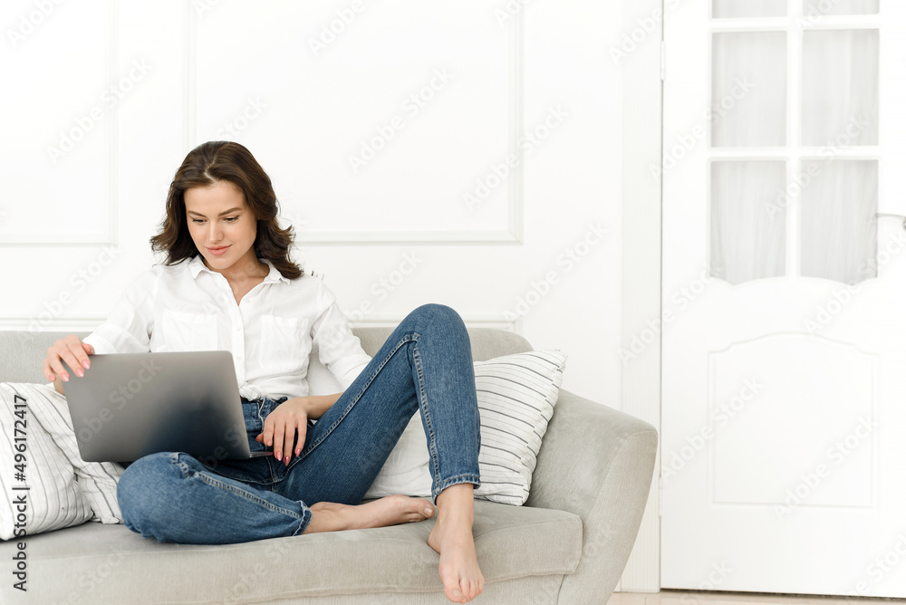 Young positive woman using laptop, checking news on internet, sitting on sofa, working remotely Young positive woman using laptop, checking news on internet, sitting on sofa, working remotely