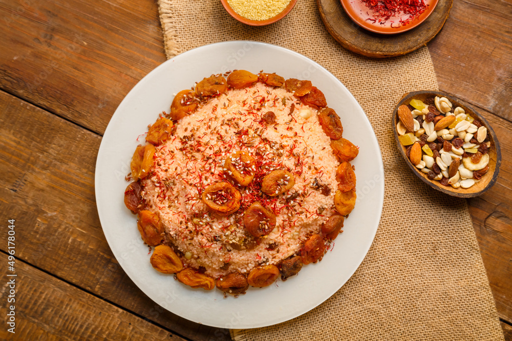 Masfouf from couscous with dried fruits in a plate on a wooden table next to saffron, and couscous groats in a bowl on a linen napkin. Masfouf from couscous with dried fruits in a plate on a wooden table next to saffron, and couscous groats in a bowl on a linen napkin.