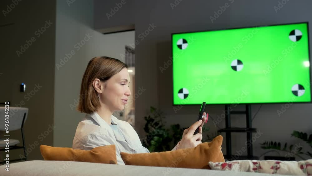Rear view the girl is sitting on the sofa in the living room with a smartphone in front of a large green screen TV. Rest at home, cozy atmosphere. Chroma key. Indoors.