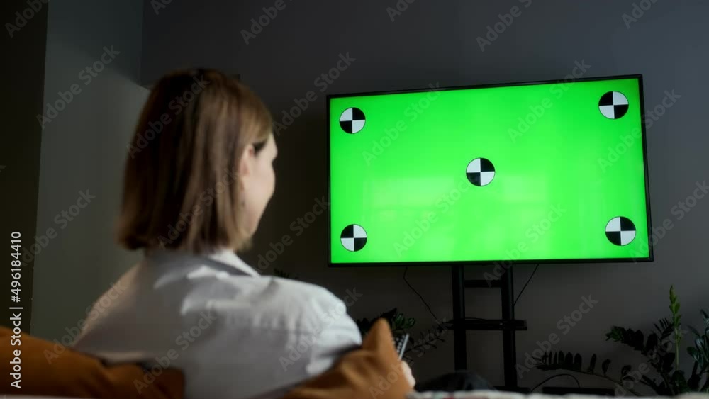A girl is sitting at home in the living room and watching TV with a green screen Chroma Key while relaxing on the sofa. Changes TV channels with the remote control. A girl is sitting at home in the living room and watching TV with a green screen Chroma Key while relaxing on the sofa. Changes TV channels with the remote control.