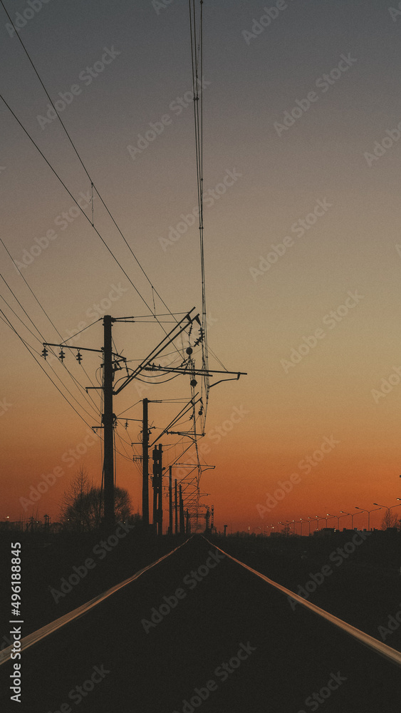 Railway tracks and power lines against the beautiful sky at sunset. Industrial landscape with railway, colorful blue sky with red clouds, sun.
