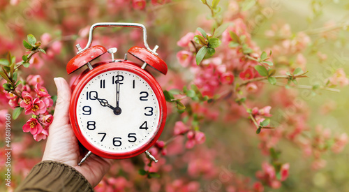 Hand holds a red classic alarm clock in a blooming flower bush. Time, spring or summer banner background.
