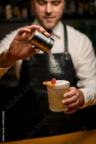 bartender gently holds glass full of foamy cocktail and sprinkles on it with sugar powder