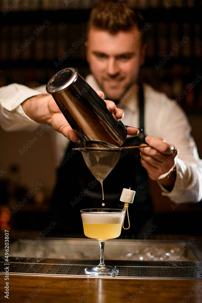 man bartender carefully filters alcoholic cocktail from steel shaker cup into glass through sieve
