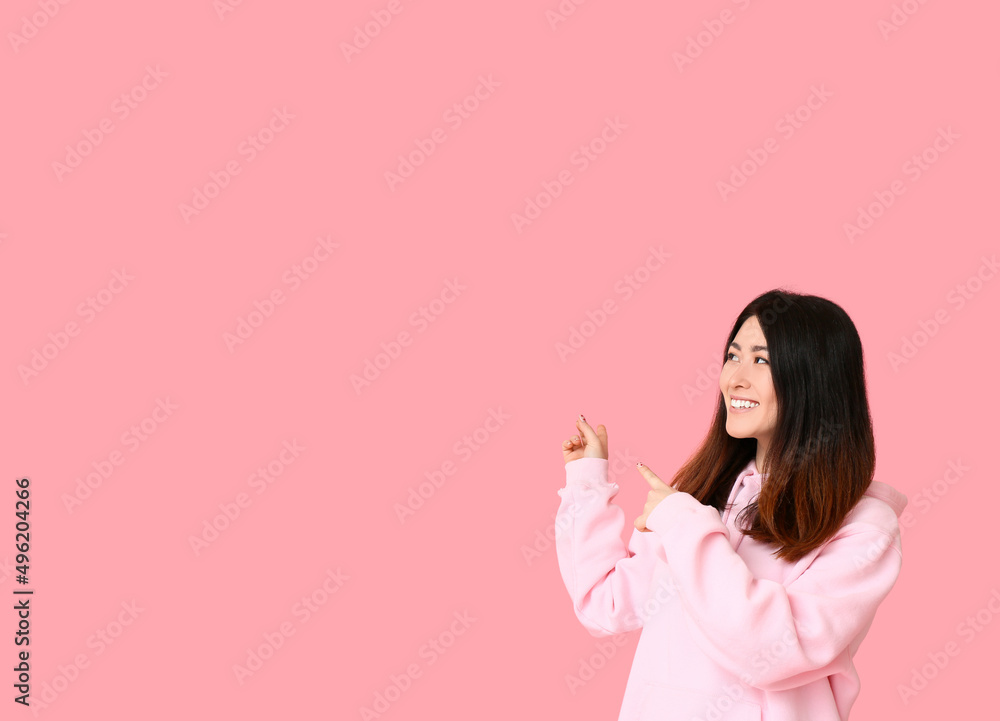 Young Asian woman in hoodie pointing at something on pink background Young Asian woman in hoodie pointing at something on pink background