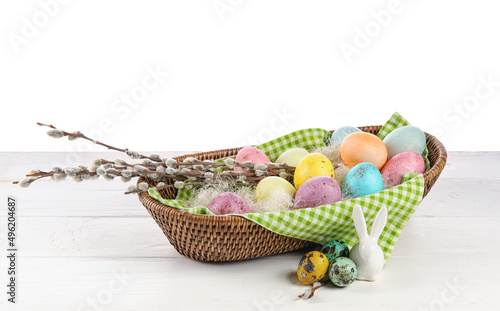 Wicker bowl with painted Easter eggs  pussy willow and figurine of bunny on table against white background