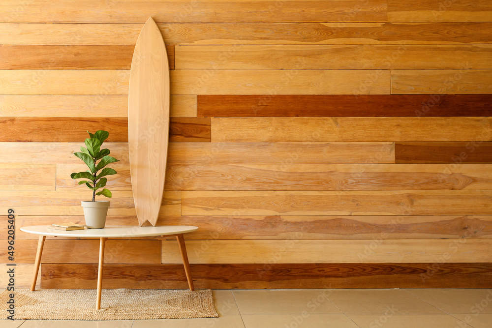 Table with surfboard, houseplant and books near wooden wall